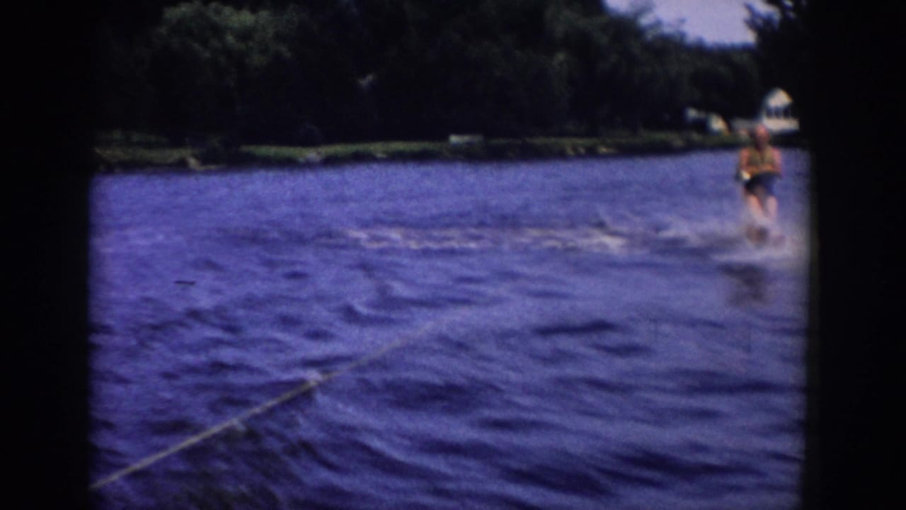 A distant skier continues across the lake in 1958 Wisconsin footage, with the tow rope, rippled water, and dark wooded shore visible.