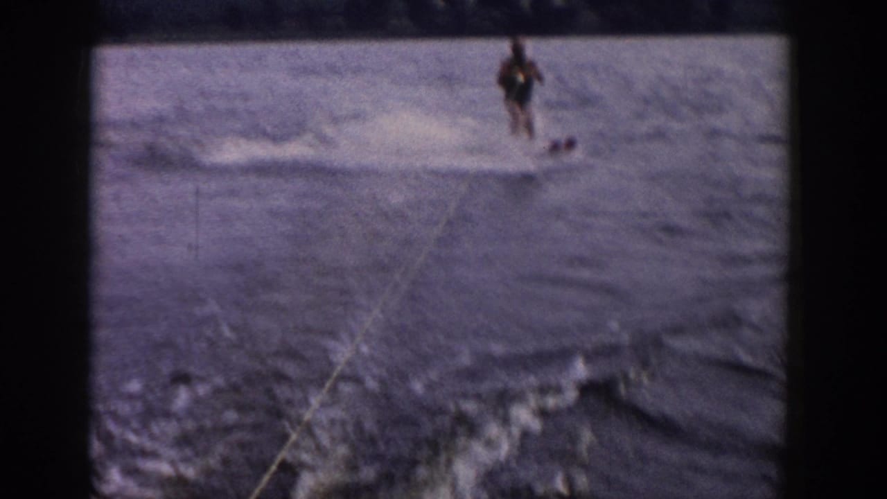 1958 Wisconsin 8mm frame showing a distant beginner water-skier on a lake, seen from a moving boat with tow rope, wake, and shore beyond.