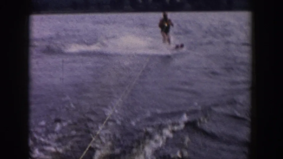 1958 Wisconsin 8mm frame showing a distant beginner water-skier on a lake, seen from a moving boat with tow rope, wake, and shore beyond.