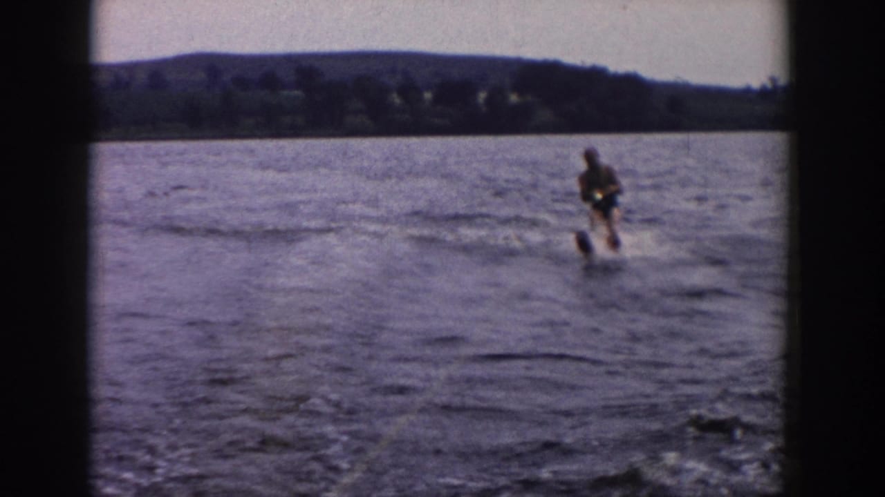 1958 Wisconsin home-movie frame of a water-skier balancing on a lake, with rippled water, spray, and a dark tree-lined shore in the distance.