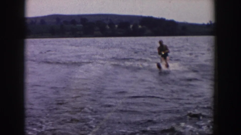 1958 Wisconsin home-movie frame of a water-skier balancing on a lake, with rippled water, spray, and a dark tree-lined shore in the distance.