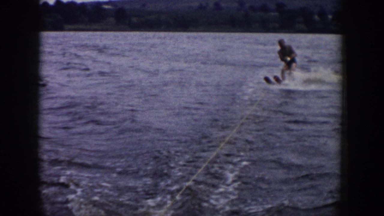 1958 Wisconsin 8mm frame showing a beginner water-skier leaning behind a boat, with yellow tow rope, splashing wake, and distant shore.