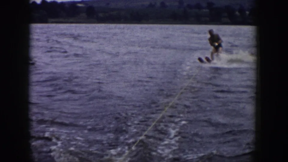 1958 Wisconsin 8mm frame showing a beginner water-skier leaning behind a boat, with yellow tow rope, splashing wake, and distant shore.