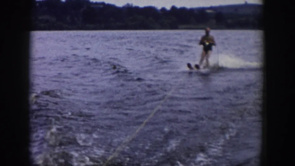 1958 Wisconsin home-movie frame of a water-skier near the edge of the shot, pulled across the lake as spray rises behind the skis.