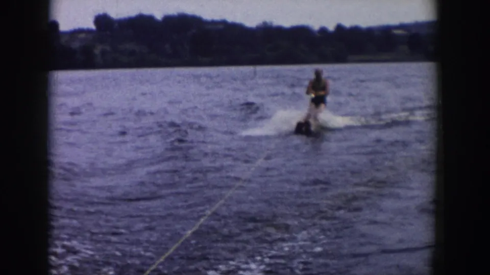 1958 Wisconsin 8mm frame showing an adult water skier behind a boat, crossing wake on a lake with trees in the distance.