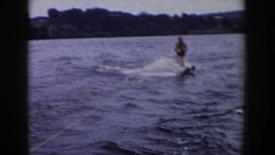 1958 Wisconsin home-movie frame of an adult skier carving across boat wake, with tow rope visible and shoreline in the background.