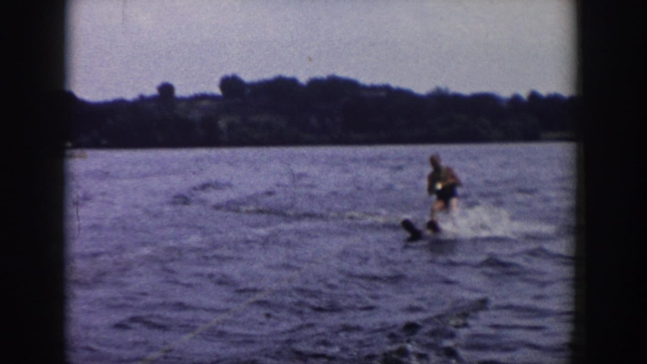 Adult water skier in 1958 Wisconsin lake footage, balancing behind a boat with spray and a wooded shore visible.