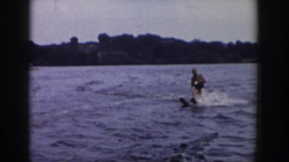 Adult water skier in 1958 Wisconsin lake footage, balancing behind a boat with spray and a wooded shore visible.