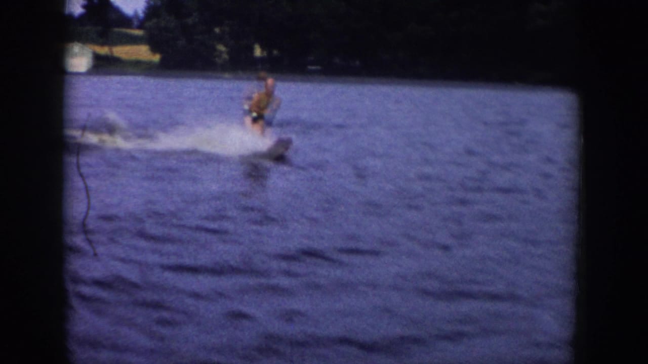 Water skier upright on a lake, leaving spray behind him near a wooded shore.