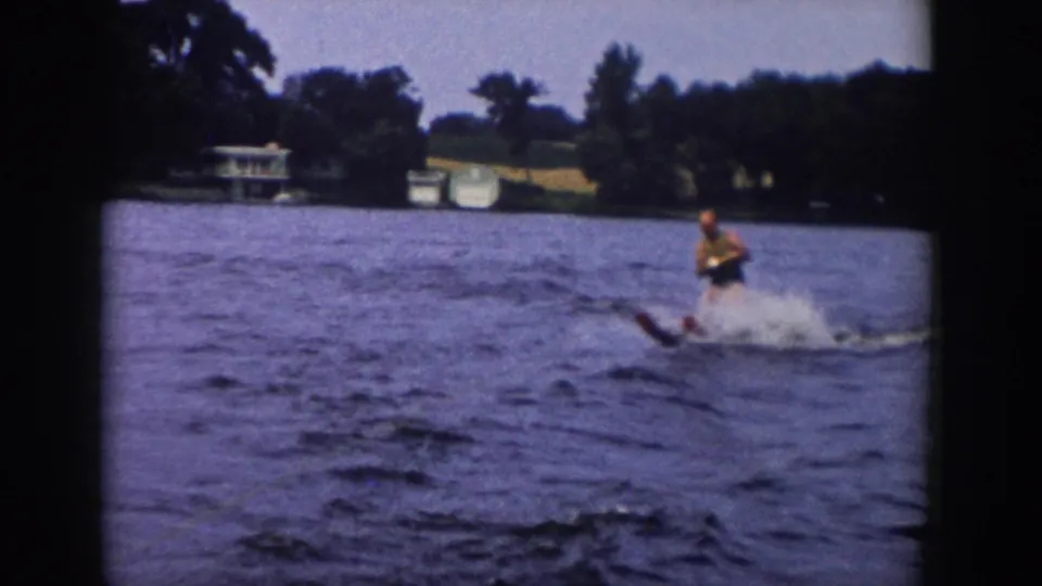 1958 Wisconsin lake footage shows the skier leaning while holding a tow rope, with distant shoreline buildings behind the water.