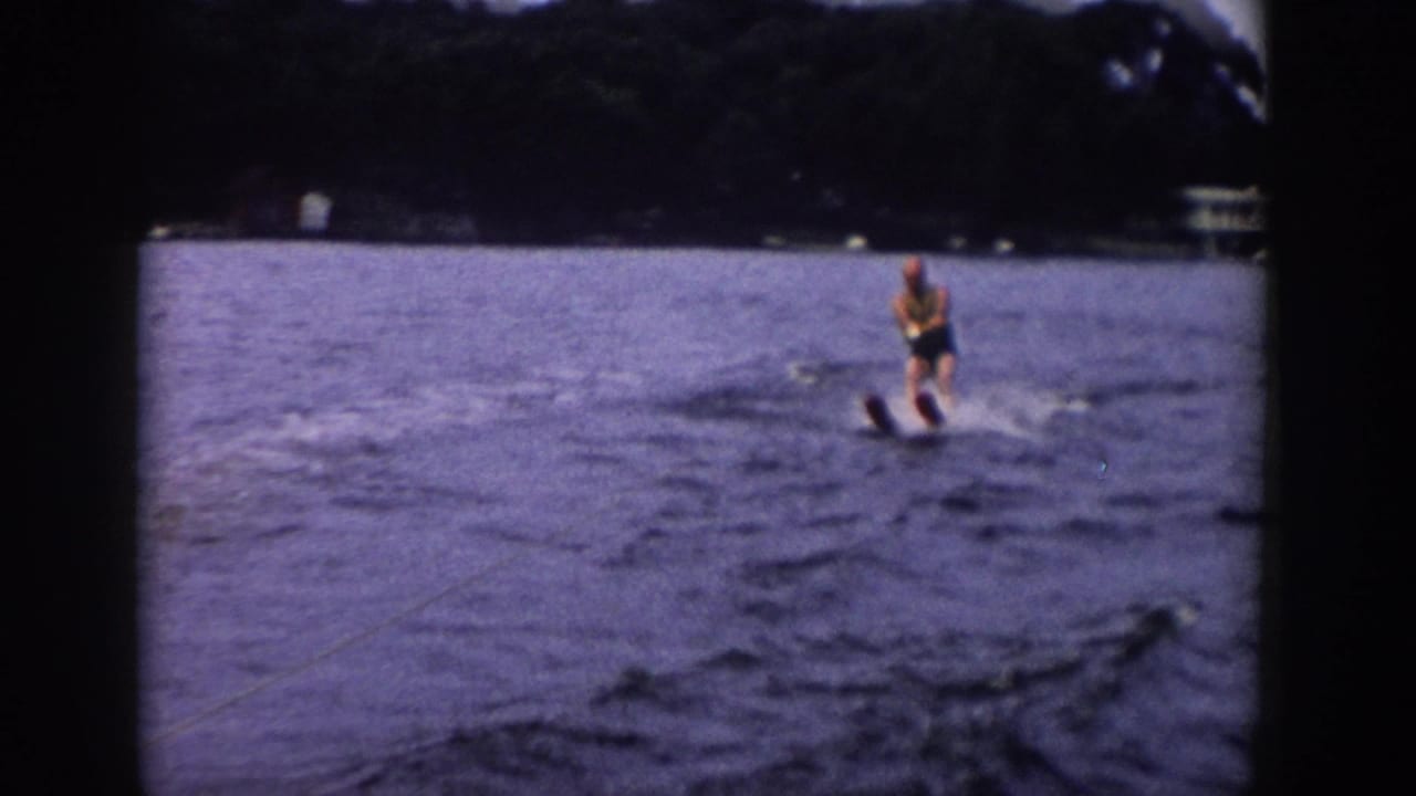1958 Wisconsin home-movie frame showing a man on waterskis at mid-distance, with open water and a dark wooded shore behind him.