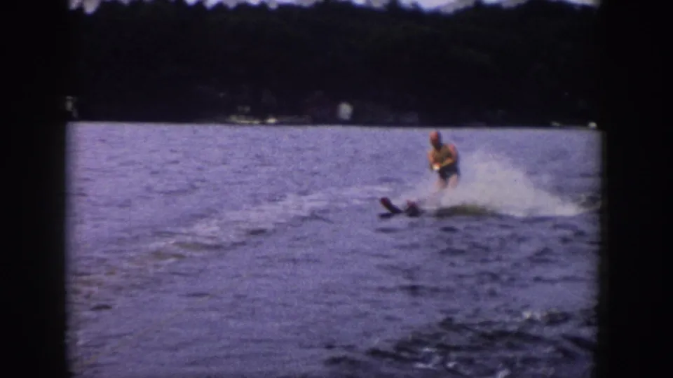 1958 Wisconsin 8mm frame showing a man water skiing across a lake, with spray behind him and dark film borders around the scene.