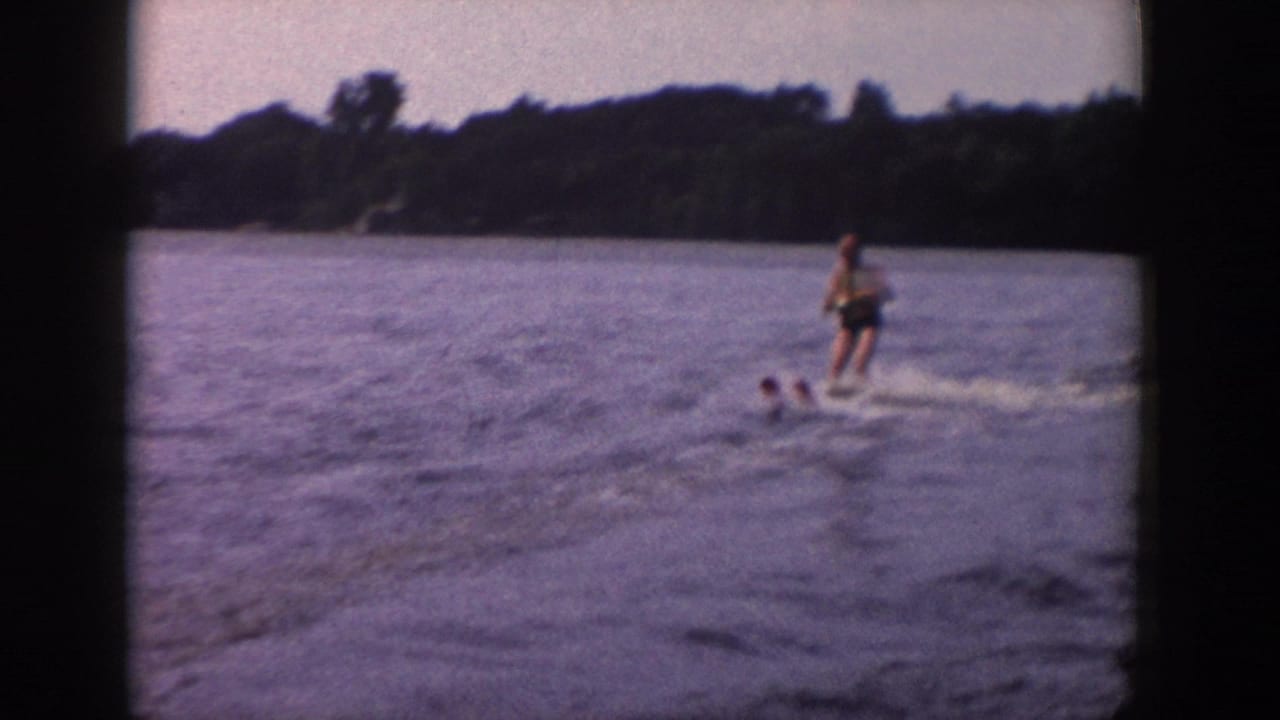 A home-movie frame shows the skier farther across the water, with a wooded shoreline and soft 8mm color fading visible.