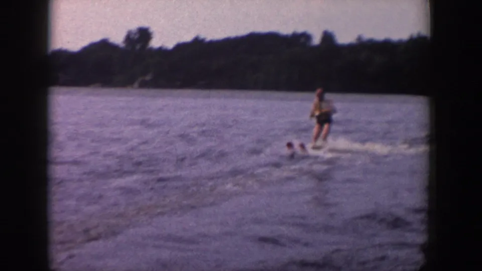 A home-movie frame shows the skier farther across the water, with a wooded shoreline and soft 8mm color fading visible.