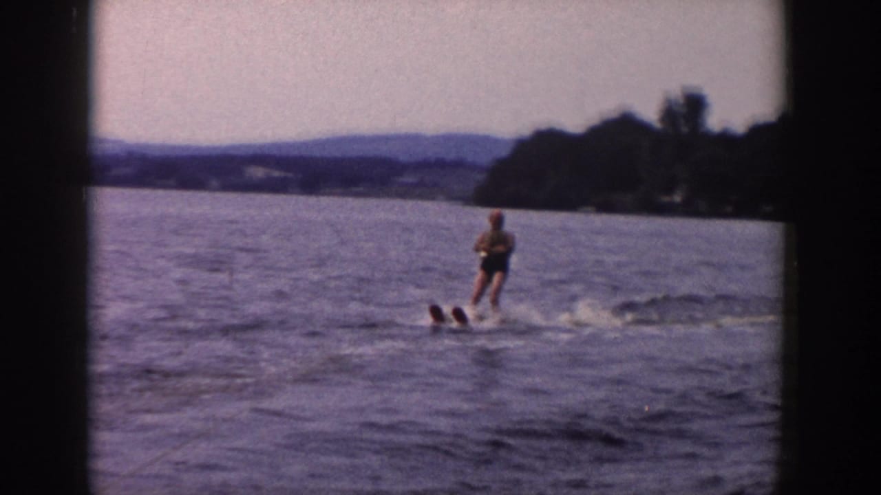 The skier crosses open water with a broad lake view, distant hills, wooded shore, and visible 8mm frame borders.