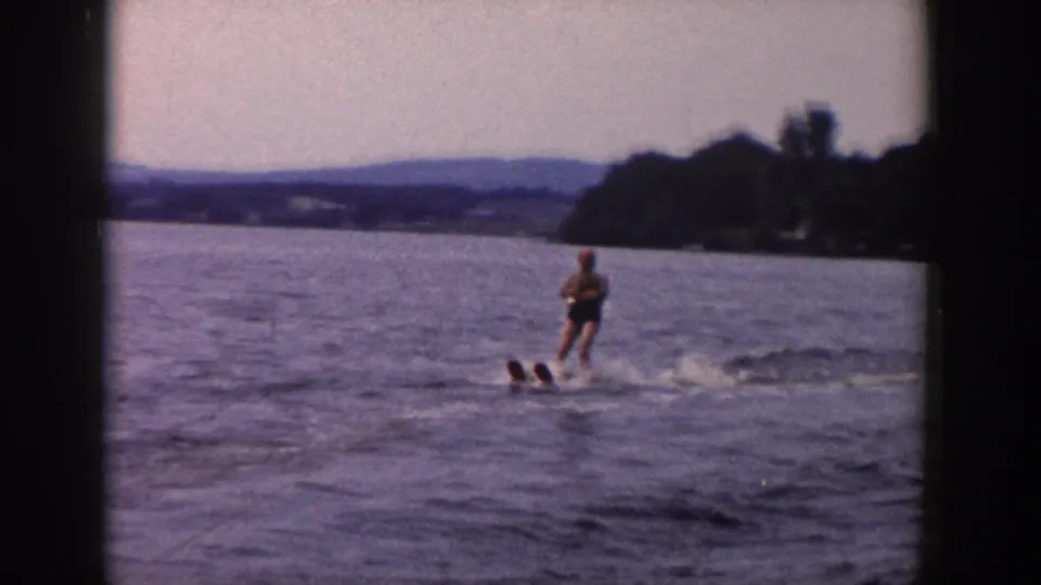 The skier crosses open water with a broad lake view, distant hills, wooded shore, and visible 8mm frame borders.