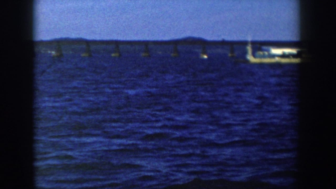 1958 Wisconsin 8mm frame showing a distant flatbed barge near a long bridge across deep blue lake water.
