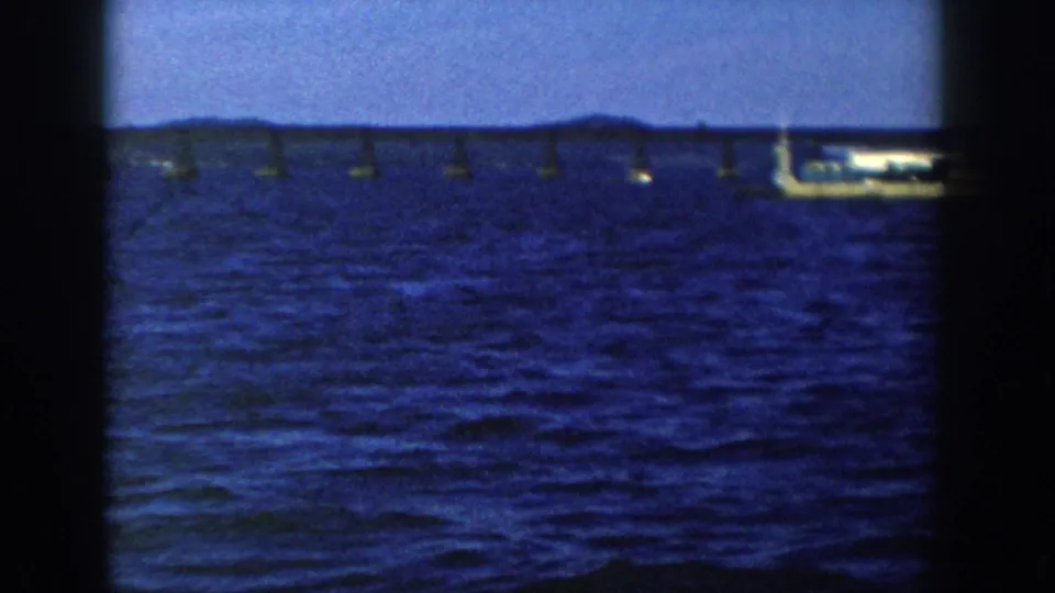 1958 Wisconsin 8mm frame showing a distant flatbed barge near a long bridge across deep blue lake water.