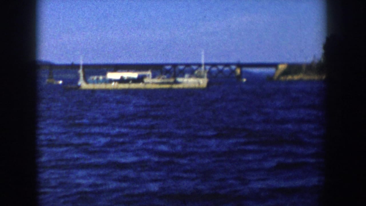 Long flatbed barge carrying a white structure passes in front of a bridge over water.