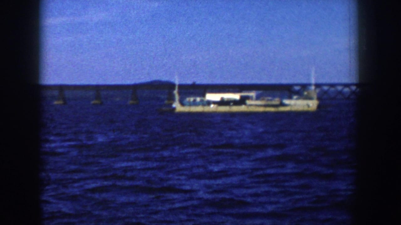 A 1958 Wisconsin 8mm frame shows the barge centered on the lake with bridge supports and low shoreline behind it.