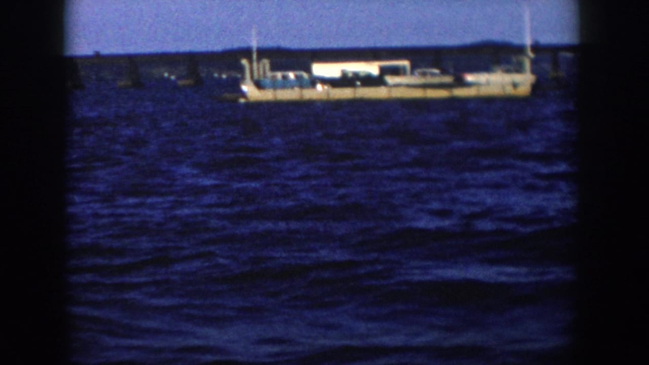 1958 Wisconsin 8mm frame showing a large light-colored ship riding on choppy blue water with a low distant shoreline behind it.