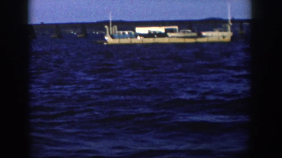 1958 Wisconsin 8mm frame showing a large light-colored ship riding on choppy blue water with a low distant shoreline behind it.
