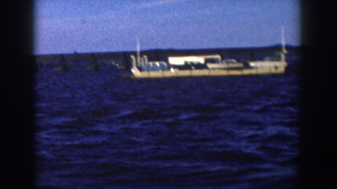 1958 Wisconsin 8mm frame showing a long barge-like vessel on blue water with a low shoreline and dark film edges.