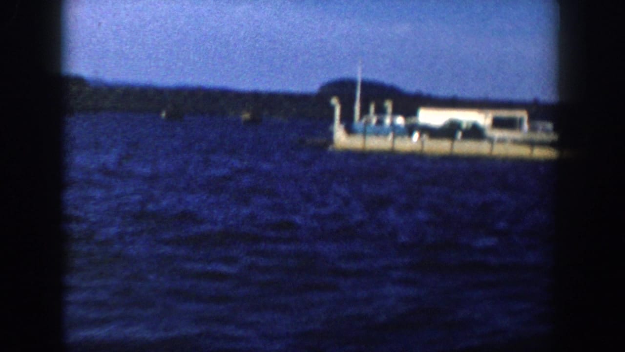1958 Wisconsin 8mm frame showing the barge-like vessel farther right, with open water, low shore, and soft handheld framing.