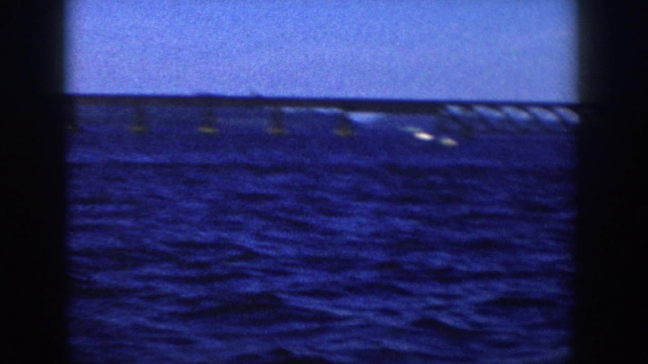 1958 Wisconsin 8mm frame showing blue lake water, clear sky, a distant bridge, and small bright wakes from a boat viewpoint.