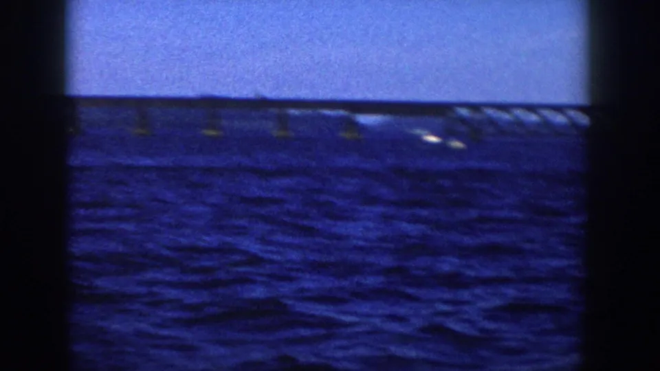 1958 Wisconsin 8mm frame showing blue lake water, clear sky, a distant bridge, and small bright wakes from a boat viewpoint.