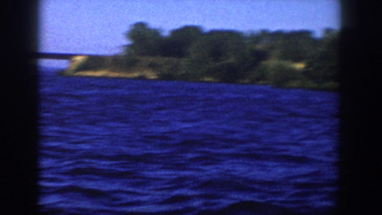 1958 Wisconsin 8mm frame from a moving boat, with open blue water, distant bridge, and wooded shoreline at the right edge.