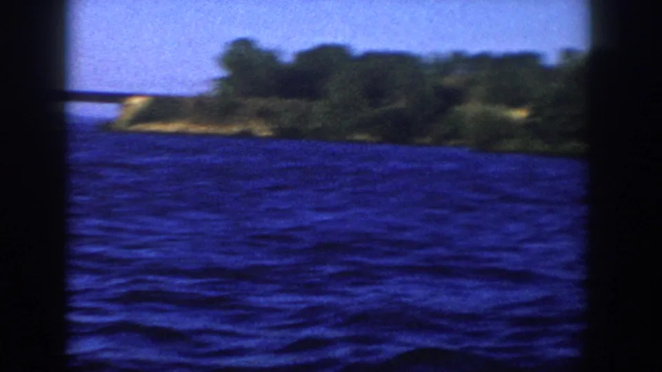 1958 Wisconsin 8mm frame from a moving boat, with open blue water, distant bridge, and wooded shoreline at the right edge.