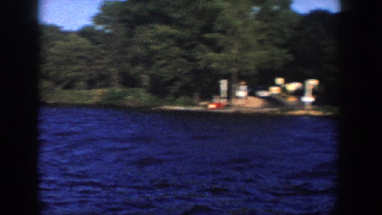 1958 Wisconsin 8mm frame showing a wooded lakeshore with a small dock area, posts, blue water, and a red buoy near shore.