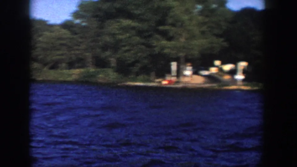 1958 Wisconsin 8mm frame showing a wooded lakeshore with a small dock area, posts, blue water, and a red buoy near shore.