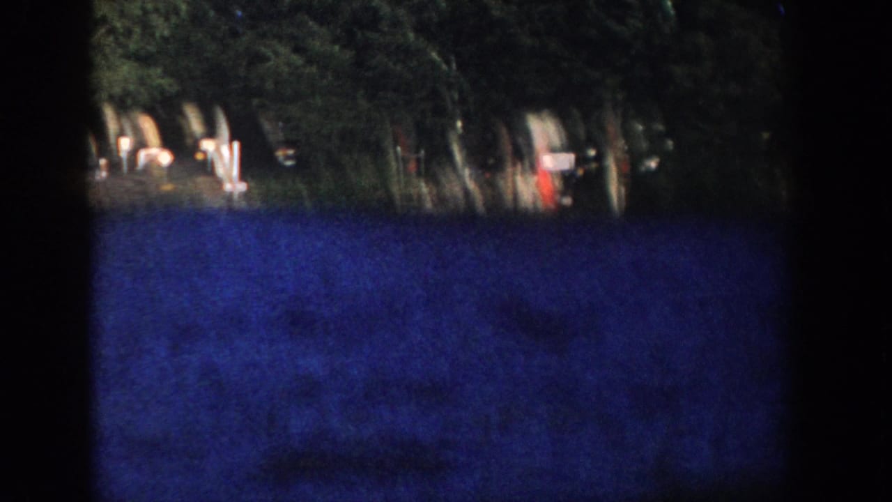 1958 Wisconsin 8mm frame of a shaded wooded lakeshore with dock posts, blue water, and small indistinct objects near the shoreline.