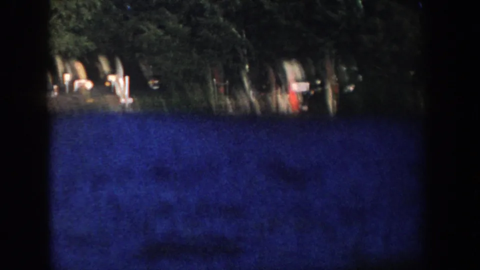 1958 Wisconsin 8mm frame of a shaded wooded lakeshore with dock posts, blue water, and small indistinct objects near the shoreline.
