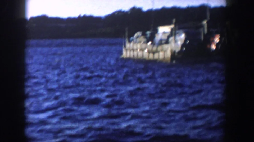 1958 Wisconsin 8mm frame with a small vehicle-laden boat crossing choppy lake water near a wooded shore.
