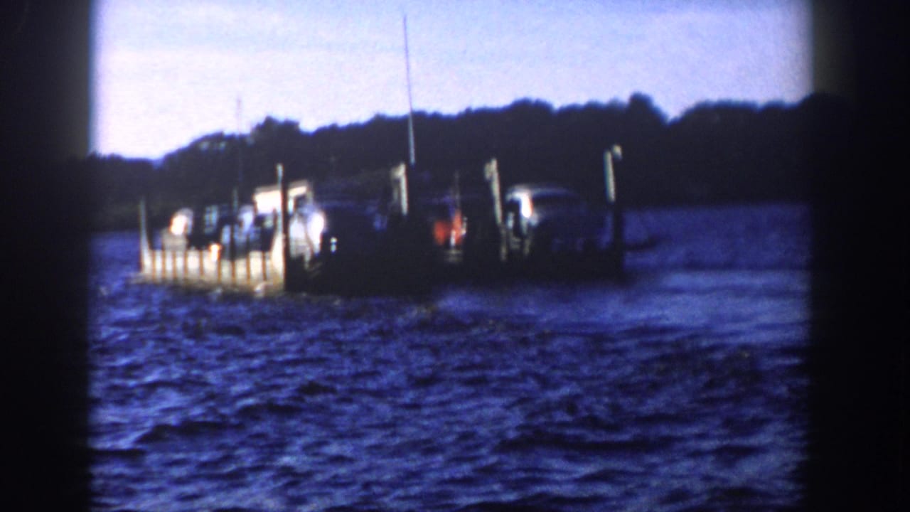 1958 Wisconsin 8mm frame showing a vehicle-laden boat near upright pilings, with dark wooded shoreline behind it.
