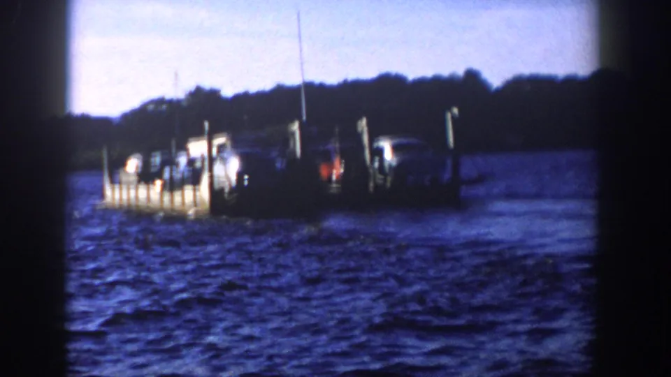 1958 Wisconsin 8mm frame showing a vehicle-laden boat near upright pilings, with dark wooded shoreline behind it.