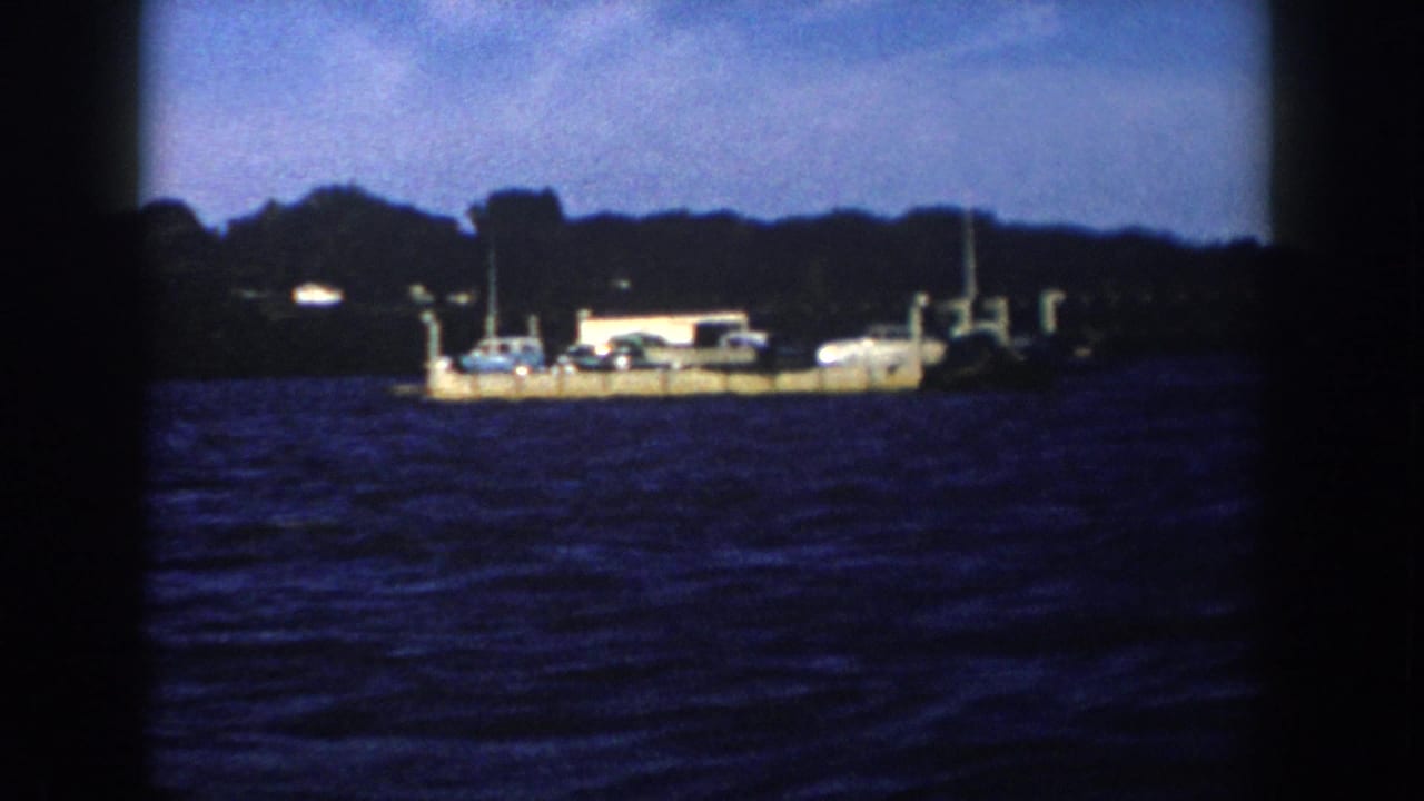 A low platform with cars and a small structure sits across dark water near a wooded shore.