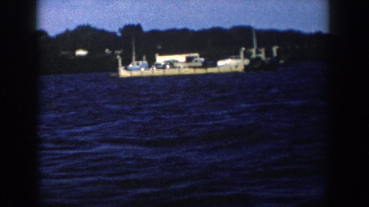 1958 Wisconsin 8mm frame of a ferry-like dock or barge carrying cars and a small structure beside a wooded shoreline.