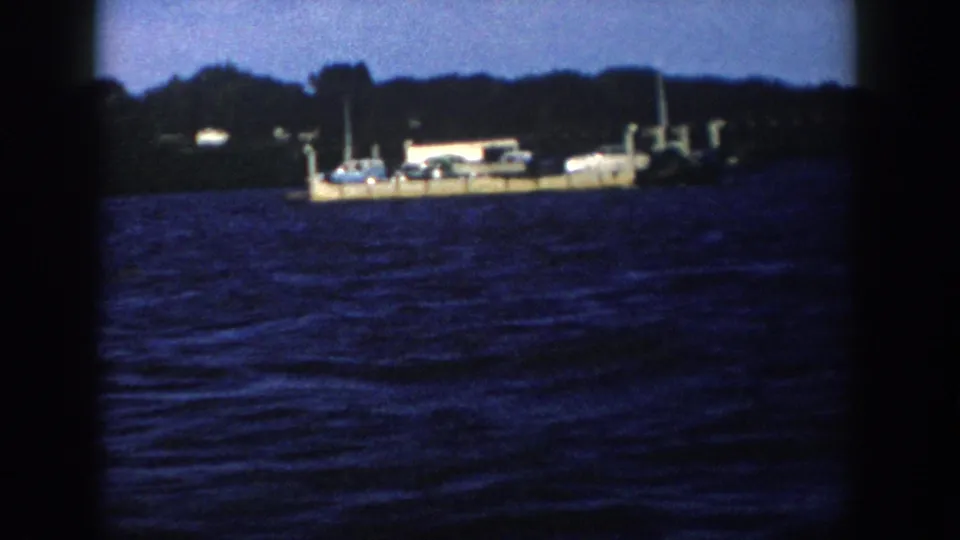 1958 Wisconsin 8mm frame of a ferry-like dock or barge carrying cars and a small structure beside a wooded shoreline.