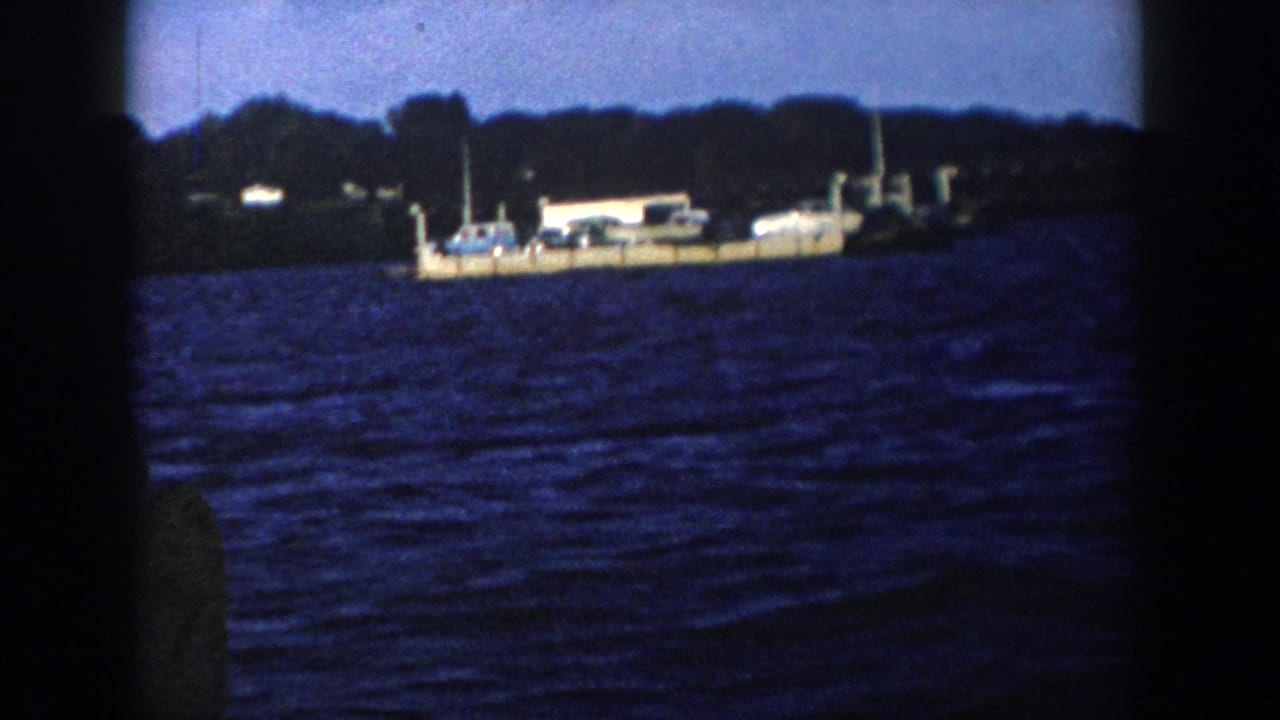 1958 Wisconsin home-movie frame showing cars on a distant dock or floating platform against trees and dark blue water.