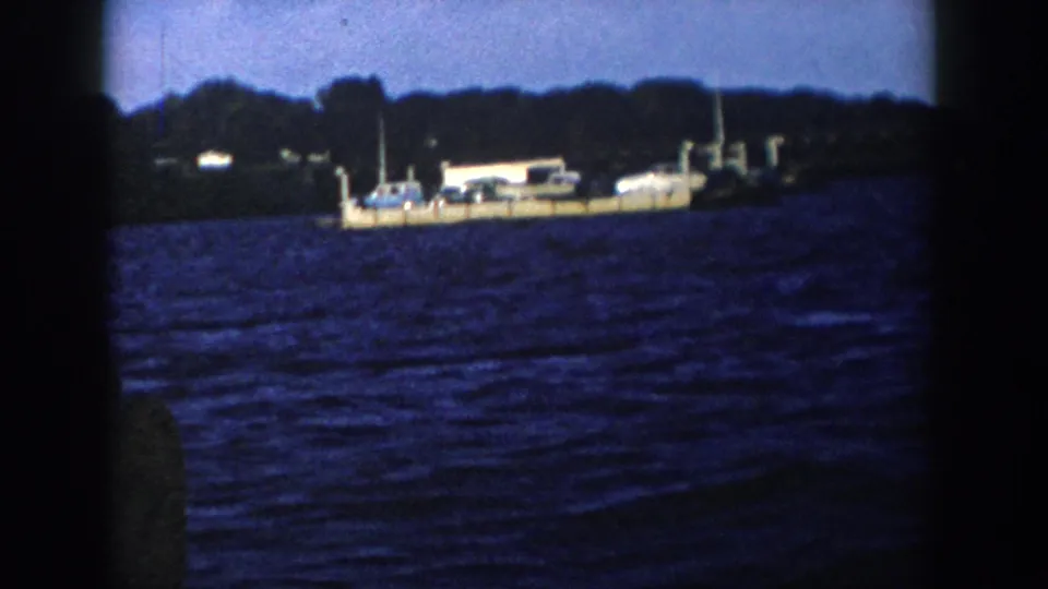 1958 Wisconsin home-movie frame showing cars on a distant dock or floating platform against trees and dark blue water.