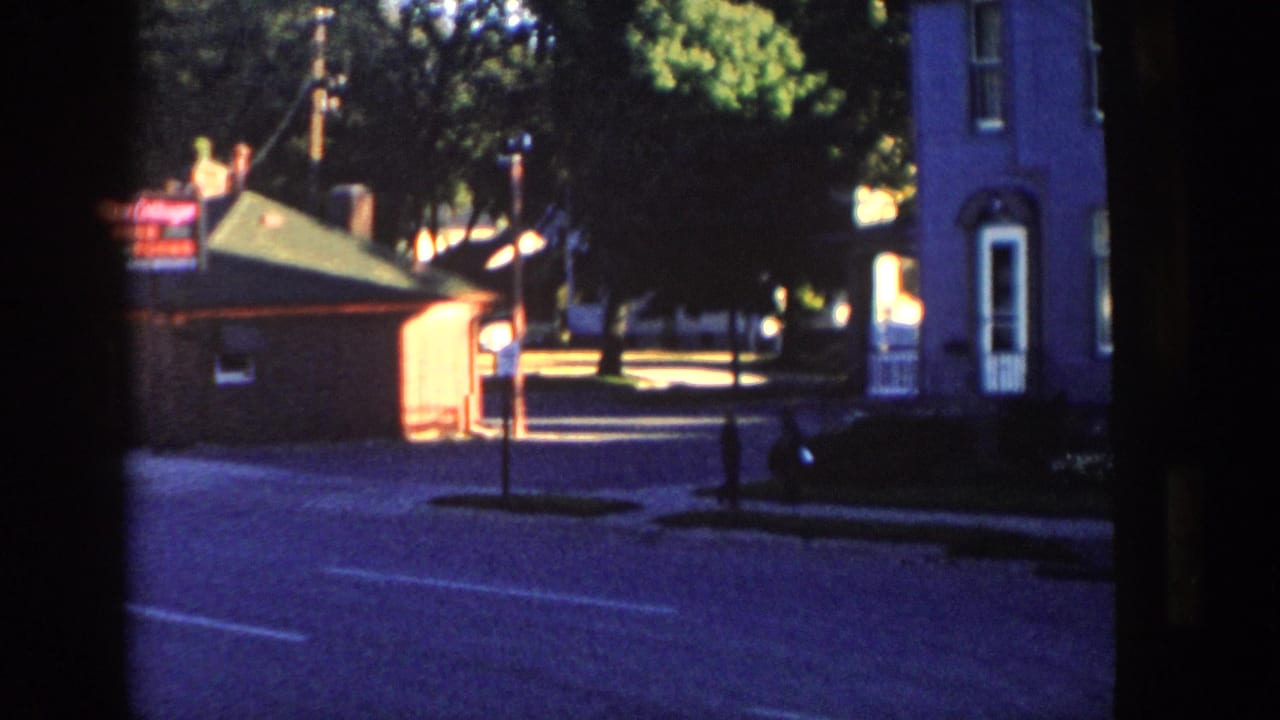 1958 Wisconsin home-movie frame of a quiet village street with a brick building, lit sign, tall house, trees, and a distant pedestrian.