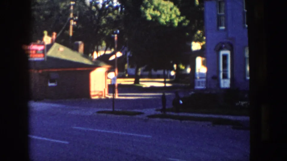 1958 Wisconsin home-movie frame of a quiet village street with a brick building, lit sign, tall house, trees, and a distant pedestrian.