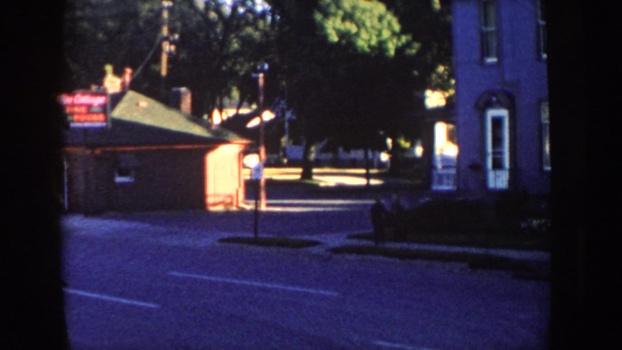Similar street view with a brick building, illuminated sign, house facade, road, sidewalk, trees, and a small pedestrian figure.