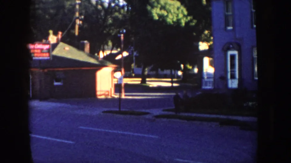 1958 Wisconsin street view showing a brick building with an illuminated sign, a tall house, tree shade, road surface, and a small pedestrian figure.