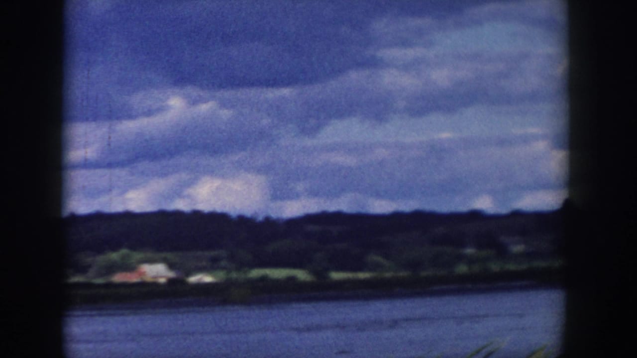Wide 1958 Wisconsin home-movie view across a lake toward small shore buildings, wooded hills, and layered clouds.
