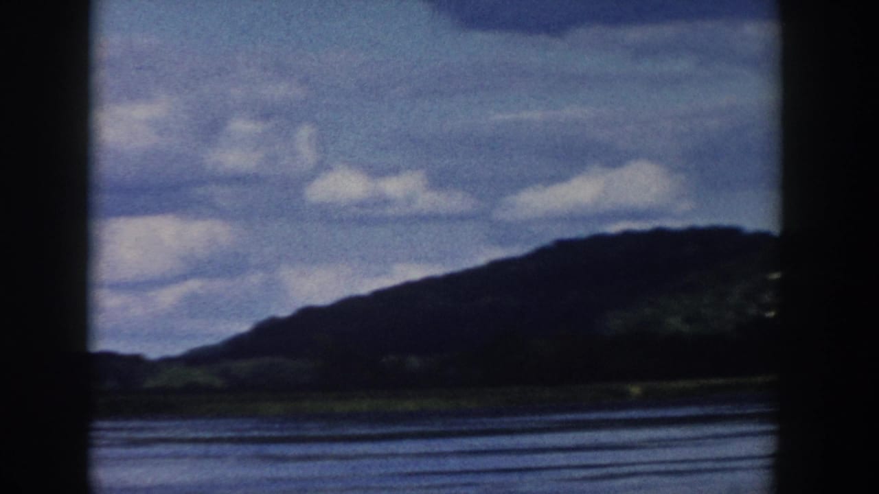 1958 Wisconsin scenic frame with a wooded hill, water in the foreground, and pale clouds across a soft blue sky.